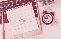 Flat lay workspace setup featuring a spiral calendar pad, small alarm clock, pen, and laptop representing scheduling a 30-minute web design discovery call.